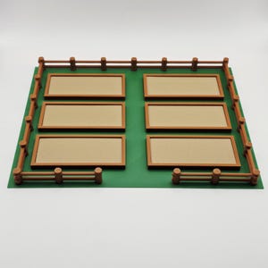 K&ouml;nnte beinhalten: Ein Miniaturmodell eines Gartenbeets mit sechs rechteckigen Pflanzbeeten. Die Beete sind beige, braun umrandet und auf einer gr&uuml;nen Basis angeordnet. Ein brauner Zaun umgibt den Garten und schafft eine detaillierte, kleine Landschaft.