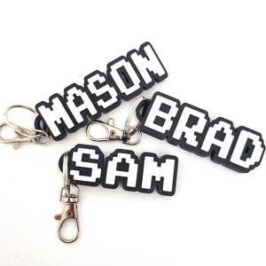 May include: Three black and white pixelated name keyrings. The names "MASON", "BRAD", and "SAM" are displayed in a block letter font. Each keyring has a silver-coloured clasp and ring.