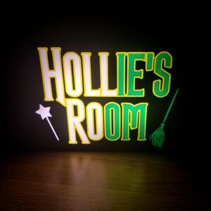 May include: A neon sign displaying the words "HOLLIE'S ROOM" in white and green, with a yellow border. A white wand and a green broom are featured. The background is black, with a wooden surface below.