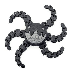 May include: A black articulated fidget toy in the shape of a smiling face with six segmented arms. The face has a grey mouth and two small white eyes. The arms are made of linked segments, allowing for flexible movement and play.