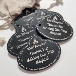 May include: Black oval tags with white text and a decorative border. The tags say "Walt Disney World" and "Thanks For Making Our Trip Magical." A small bag is visible in the background.
