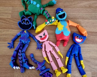 May include: A selection of brightly coloured, articulated character toys. The toys include a green frog-like figure, a blue cat-like figure with a crescent moon, and several figures with smiling faces in pink and blue. The toys are on a wooden surface.