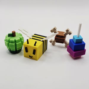 K&ouml;nnte beinhalten: Eine Sammlung farbenfroher, pixeliger Spielzeuge, darunter ein gr&uuml;ner Apfel, eine gelb-schwarze Biene mit wei&szlig;en Fl&uuml;geln, ein braun-beiges kreuzf&ouml;rmiges Objekt und ein Stapel blauer, lila und t&uuml;rkisfarbener Bl&ouml;cke mit einem wei&szlig;en Stab.