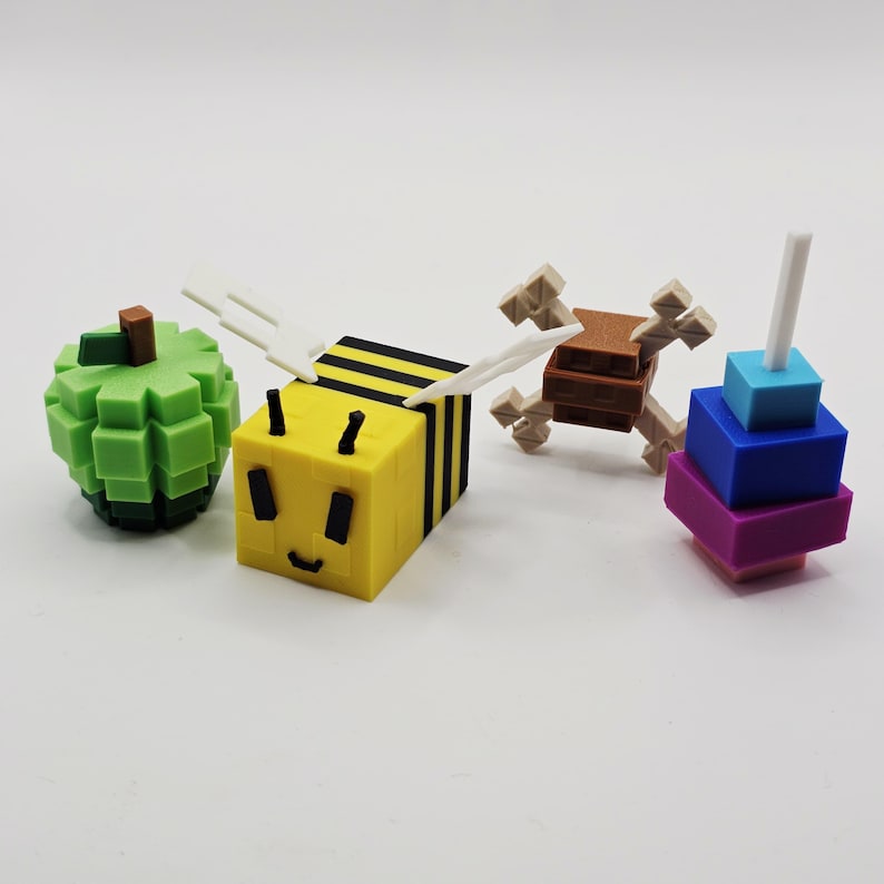 K&ouml;nnte beinhalten: Eine Sammlung farbenfroher, pixeliger Spielzeuge, darunter ein gr&uuml;ner Apfel, eine gelb-schwarze Biene mit wei&szlig;en Fl&uuml;geln, ein braun-beiges kreuzf&ouml;rmiges Objekt und ein Stapel blauer, lila und t&uuml;rkisfarbener Bl&ouml;cke mit einem wei&szlig;en Stab.
