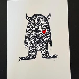 May include: A black and white illustration of a friendly monster with a red heart on a white card. The monster has horns, a single tooth, and is covered in white dots. The card is on a black surface.