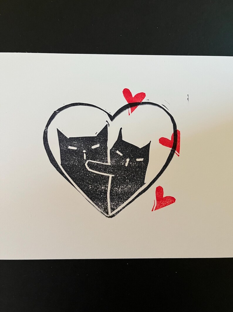 May include: Two black cats are inside a heart shape with red hearts around it. The cats are facing each other and appear to be hugging.