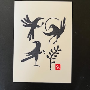 May include: A cream-colored card features a black linocut print of three stylized birds and a sprig of leaves. A small red square with a symbol is in the lower right corner. The birds are in various poses, with one holding a small object.