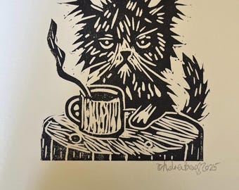 May include: Black and white linocut print of a grumpy cat with a coffee mug on a wooden surface. The cat has spiky fur and a stern expression. Steam rises from the mug. Signed "Andrea Krug 2025".