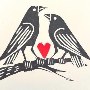 May include: Two black birds perched on a branch with a red heart between them. The birds are facing each other and have a simple, stylized design.