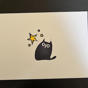 Puede incluir: Un gato negro con ojos blancos está sentado mirando hacia arriba a una estrella amarilla con un halo amarillo. Hay pequeños círculos amarillos alrededor de la estrella.