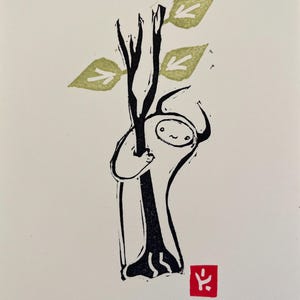 May include: A black and white illustration of a small, white creature hugging a tree with green leaves. The creature has a simple, smiling face. A red stamp with a black symbol is in the bottom right corner.