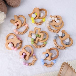 Puede incluir: Una colección de mordedores de madera para bebés en varias formas, incluyendo estrellas, pájaros y cruces. Cada mordedor presenta un anillo de madera, cuentas de silicona y una bola tejida. Los nombres de los bebés están escritos con cuentas de letras de madera. Los colores incluyen rosa, amarillo, verde y azul.