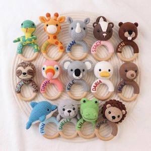 May include: A collection of colorful, crocheted animal baby rattles. Each rattle features a different animal design, including a snake, giraffe, rhino, skunk, bear, sloth, parrot, koala, duck, bear, dolphin, seal, frog, and lion. The names are embroidered on the wooden ring.