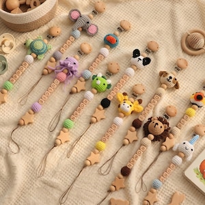 Op de afbeelding: Een verzameling houten fopspeenclips met verschillende gehaakte dierenontwerpen, waaronder een schildpad, olifant en leeuw. Elke clip heeft een houten ster en gepersonaliseerde naamkralen. De clips liggen op een crèmekleurig, getextureerd oppervlak.