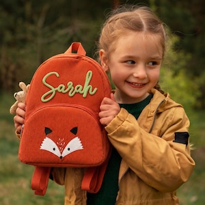 Puede incluir: Una mochila de pana color óxido con un diseño de cara de zorro en el bolsillo delantero y el nombre Sarah bordado en letra cursiva verde claro. La mochila tiene un asa superior, correas ajustables para los hombros y un pequeño juguete tejido.