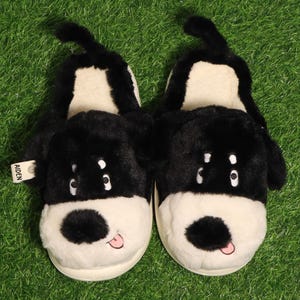 Puede incluir: Un par de zapatillas con forma de perro en blanco y negro con una textura esponjosa. Las zapatillas tienen una base blanca, orejas negras y una cola negra. La cara del perro presenta una nariz negra, ojos blancos y una lengua rosa. La etiqueta dice "AIDEN".