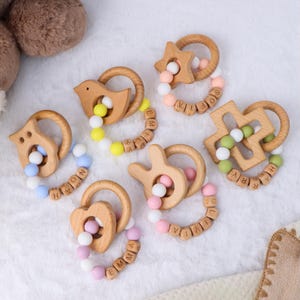 Puede incluir: Una colección de mordedores de bebé de madera en varias formas, incluyendo un pájaro, una estrella y un corazón. Cada mordedor tiene un anillo de madera y cuentas de silicona de colores, algunas con nombres grabados. Los mordedores están diseñados para bebés.