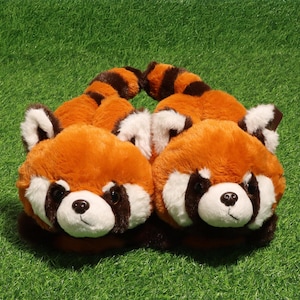 Può includere: Un paio di pantofole in peluche a forma di panda rosso, con morbida pelliccia arancione e bianca, macchie marrone scuro intorno agli occhi, naso nero e code a strisce sul retro. Le pantofole presentano orecchie arrotondate e una texture soffice.