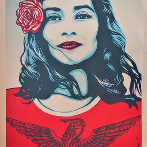 Peut inclure: Affiche représentant une femme aux cheveux bleus et une rose rouge. Elle porte un haut rouge avec un aigle. Le texte "WE THE PEOPLE" et "DEFEND DIGNITY" est en bas de l'affiche.