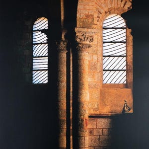 May include: Architectural photograph of a stone church interior with arched windows. The windows have white and black diagonal lines. The walls are made of light brown bricks. The image is dark with sunlight illuminating the windows and columns.