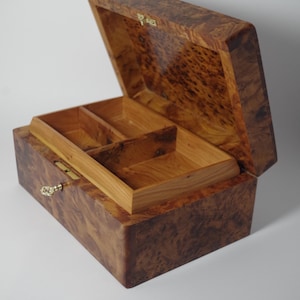 May include: A wooden jewelry box with a hinged lid, open to reveal a tray with compartments. The box is made of burl wood, with a natural grain pattern and a warm, brown color. The box has a gold-colored clasp and hinges.