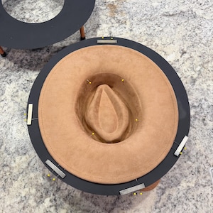 May include: A tan-colored fedora hat is placed inside a dark gray hat-shaping tool. The hat is secured with yellow pins. The hat has a rounded crown and a wide brim. The shaping tool has a circular design.