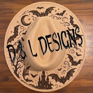 May include: A tan wide-brimmed hat featuring a dark brown printed design of bats, a crescent moon, a castle, and bare trees. The text D & L DESIGNS is printed in black across the center of the hat, which has a structured crown and flat brim.