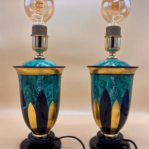 May include: A pair of table lamps featuring a green malachite-patterned body with gold petal-shaped accents and a black base. Each lamp has a gold-toned socket and a clear glass light bulb with a visible orange filament, connected by a black cord.