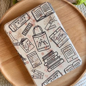 May include: A rectangular e-reader case featuring a cream-colored background with black line art illustrations of books, a tote bag labeled Reader Era, a key, bookmarks, and book-related text including Book Lovers Club and Bookworm.