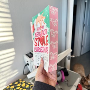 May include: A book titled "How My Neighbor Stole Christmas" by Meghan Quinn. The book has a teal cover with a Santa Claus hand illustration and a pink spine decorated with candy cane bows. The book is held in front of a blurred background.