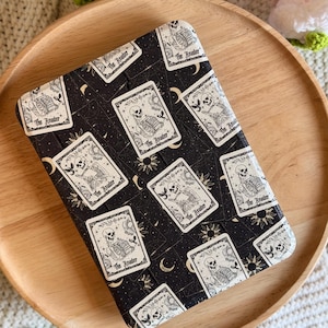 May include: A rectangular e-reader case featuring a black background with a repeating pattern of tarot cards. Each card displays a skeleton reading a book with the text The Reader, surrounded by celestial motifs like suns and crescent moons.