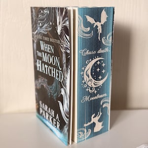 May include: A hardcover book titled When the Moon Hatched by Sarah A. Parker with a black cover featuring white dragon illustrations. The page edges are decorated with a teal blue design showing silhouettes of dragons, a crescent moon, and stars.