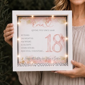 May include: A white square shadow box frame with internal LED lights containing a personalized print for an 18th birthday. The design features pink watercolor accents, the name Evin, and text detailing 18 years, 216 months, 939 weeks, 6,574 days, 157,785 hours, and a pink ribbon.