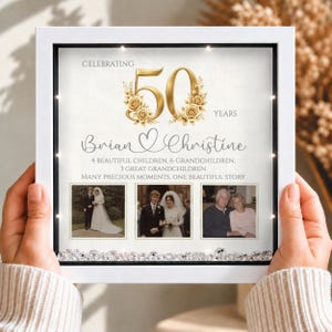 Pode incluir: Uma moldura quadrada branca tipo caixa com luzes LED integradas, apresentando um design de 50º aniversário. Exibe o texto CELEBRATING 50 YEARS, os nomes Brian e Christine, detalhes familiares e três fotografias, com cristais decorativos na base.