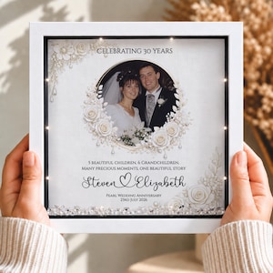 Puede incluir: Un marco cuadrado blanco tipo caja con una foto de boda, rodeada de ilustraciones florales blancas y cristales. El texto dice: CELEBRATING 30 YEARS, 5 BEAUTIFUL CHILDREN, 6 GRANDCHILDREN, MANY PRECIOUS MOMENTS, ONE BEAUTIFUL STORY, Steven and Elizabeth, PEARL WEDDING ANNIVERSARY 23RD JULY 2026.