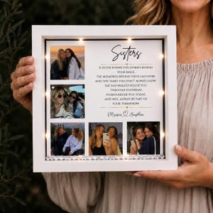 May include: A white square shadow box frame with integrated LED lights containing five photographs of two women and a printed poem titled Sisters. The text reads: A sister knows the stories behind your smile, the memories behind your laughter and the tears you don't always show. She has walked beside you through yesterday, stands beside you today, and will always be part of your tomorrow. The names Maisie and Sophie are printed at the bottom.
