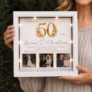 May include: A white square shadow box frame with internal LED lights, featuring a gold 50th anniversary graphic, the names Brian and Christine, and text detailing family history. Three square photos are displayed above clear crystal accents.