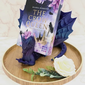 Könnte beinhalten: Ein dunkelblauer Drachen-Buchhalter hält ein Buch mit dem Titel "The Crow Queen" mit lila floralen Akzenten. Der Drache befindet sich auf einem Holztablett mit einer weißen Rose und Grünzeug.