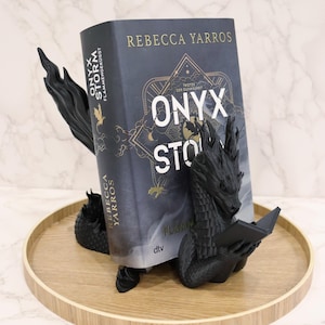 Può includere: Un fermalibri a forma di drago nero tiene un libro intitolato "Onyx Storm" di Rebecca Yarros. Il fermalibri è su un vassoio di legno. Il libro ha una copertina scura con scritte dorate e un design decorativo.