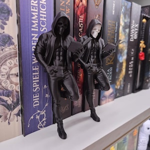 Könnte beinhalten: Zwei schwarze Figuren von Figuren mit Totenkopfmasken, Kapuzen und Jacken, die sich an ein Bücherregal lehnen. Jede Figur hält ein Buch. Die Figuren sind vor einer Reihe von Büchern mit sichtbaren Titeln positioniert.