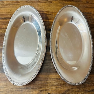 May include: Two matching oval silver-toned serving platters with a polished, reflective surface and a decorative embossed floral pattern along the outer rim. These metallic dishes feature a smooth, concave center and a raised, textured border.
