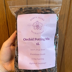 May include: A transparent bag containing orchid potting mix, with a pink label. The label reads "DARCY JAYNE Orchid Care" and "Orchid Potting Mix 1L". Additional text includes "100% Peat Free", "Premium Grade", "Long Lasting", and "Optimal Drainage".