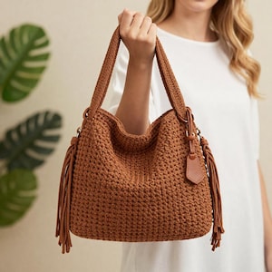 May include: A brown crochet handbag with long handles and a shoulder strap. The bag features a textured stitch pattern, a leather tag, and decorative tassels. The bag is held up by a person.