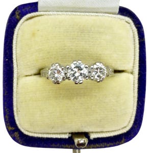 Peut inclure: Une bague en diamant à trois pierres dans un écrin en velours bleu. La bague présente trois diamants ronds sertis sur une monture en argent. L'écrin a un intérieur jaune clair et une fermeture à charnière.