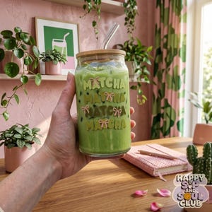 Op de afbeelding: Een doorzichtig, blikvormig glas gevuld met een felgroene matcha-drank. Het glas is bedrukt met het woord MATCHA in herhalende rijen met kleine roze strikillustraties. Het bevat een bamboe deksel en een glazen rietje.