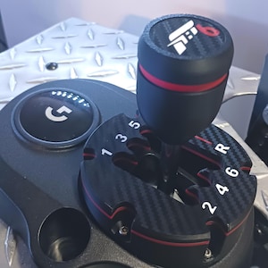 May include: A black gaming gear shifter featuring a carbon fiber pattern plate with white gear numbers 1, 3, 5, 2, 4, 6 and R. The shifter knob has a red ring and a top cap with a white logo and the number 6. A circular black button with a white G logo.