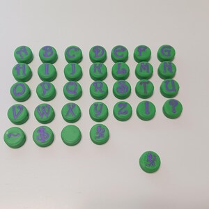 May include: Green circular objects with purple letters arranged in rows. The letters of the alphabet are printed on the green circles. The objects are on a white surface.