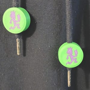 May include: Two green and purple drawstring cord ends. Each end features a green disc with a purple graphic of a running figure. The cords are black and the metal aglets are silver.
