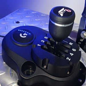 May include: A black gaming gear shifter featuring a six-speed manual pattern with reverse, marked with white numbers 1, 2, 3, 4, 5, 6, and R. The top of the shifter knob displays a pink and white logo, and a circular G logo is visible on the base.