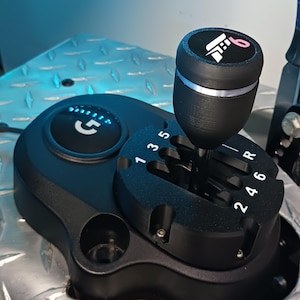 May include: Black gear shift knob with a white and pink graphic on top. The shift pattern is visible with numbers 1 through 6, plus R for reverse. The base has the letter "G" illuminated in blue.
